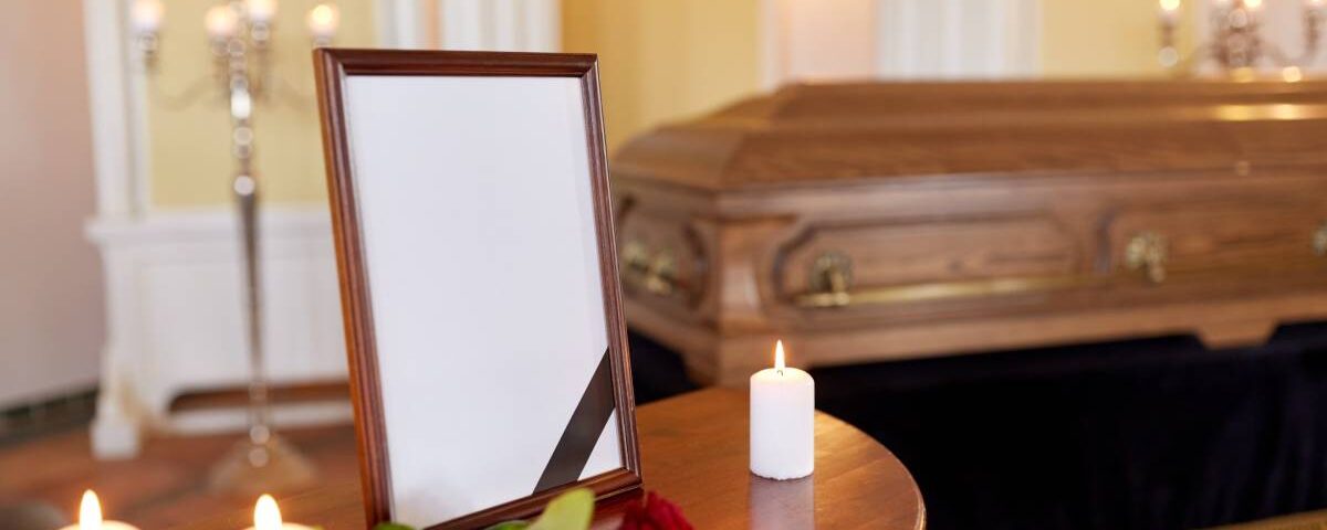 A small table displays a picture frame, lit white candles, and a rose in front of a wooden casket at a funeral.