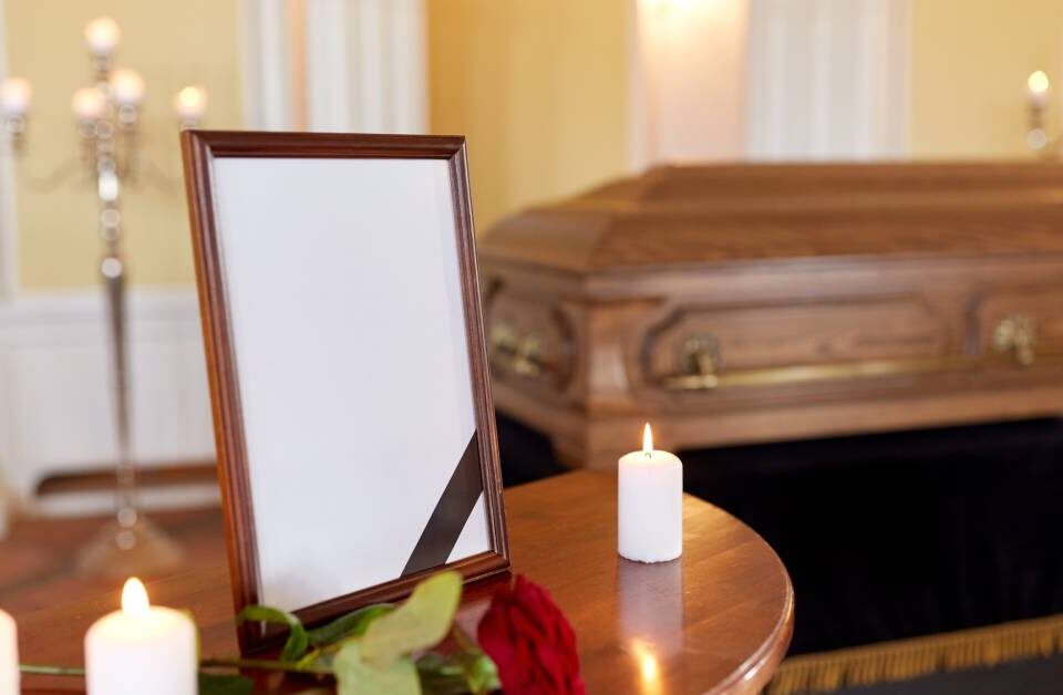 A small table displays a picture frame, lit white candles, and a rose in front of a wooden casket at a funeral.