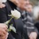 A line of people standing and wearing dark clothes at a memorial service. A man holds a white rose in the foreground.