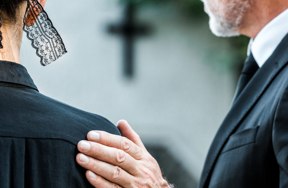 Two funeral guests face away from view. The older man places his hand on the woman's shoulder next to him.
