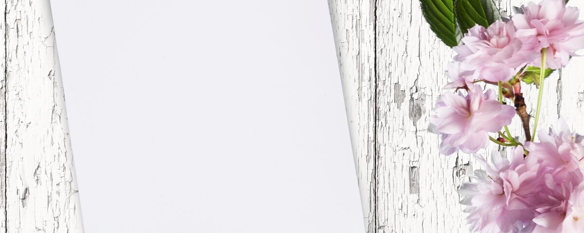 A blank piece of paper on a white, distressed wood background with pink flowers and a black ribbon to the side.