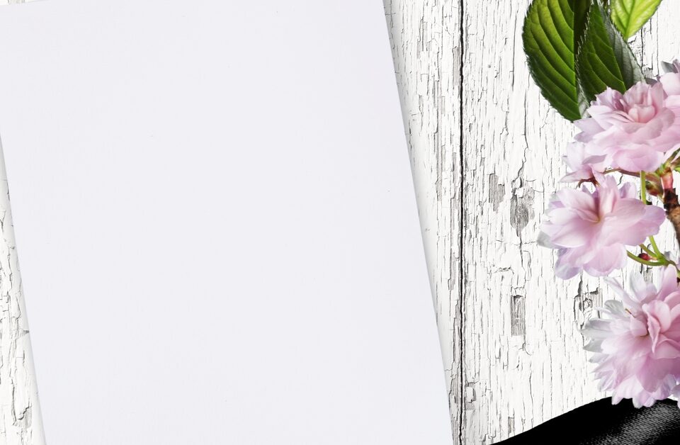 A blank piece of paper on a white, distressed wood background with pink flowers and a black ribbon to the side.