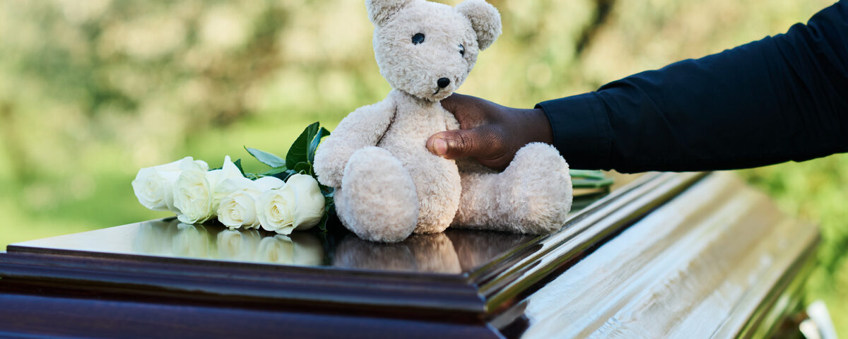 A man's arm in a black, long-sleeve shirt places an off-white teddy bear on a casket next to four white roses.