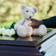 A man's arm in a black, long-sleeve shirt places an off-white teddy bear on a casket next to four white roses.
