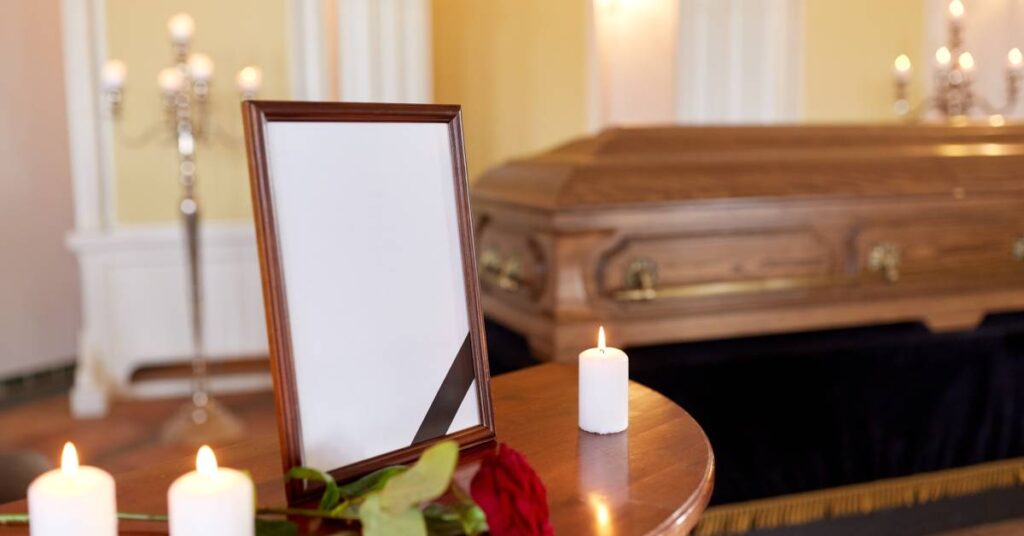 A small table displays a picture frame, lit white candles, and a rose in front of a wooden casket at a funeral.