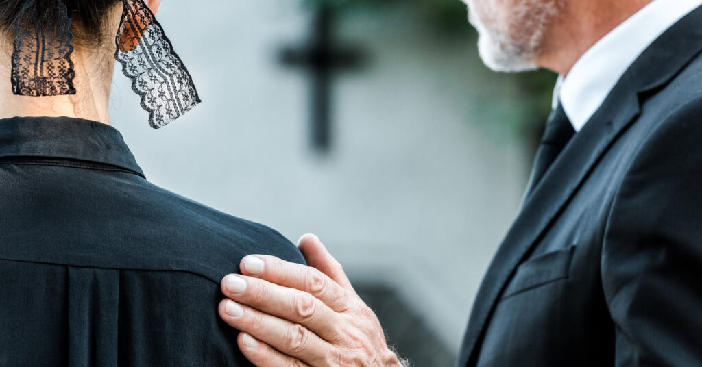 Two funeral guests face away from view. The older man places his hand on the woman's shoulder next to him.