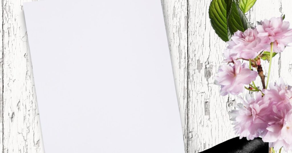 A blank piece of paper on a white, distressed wood background with pink flowers and a black ribbon to the side.